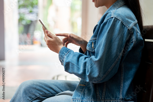 A close-up of a woman in a denim jacket using her smartphone while sitting on a bench in the city.