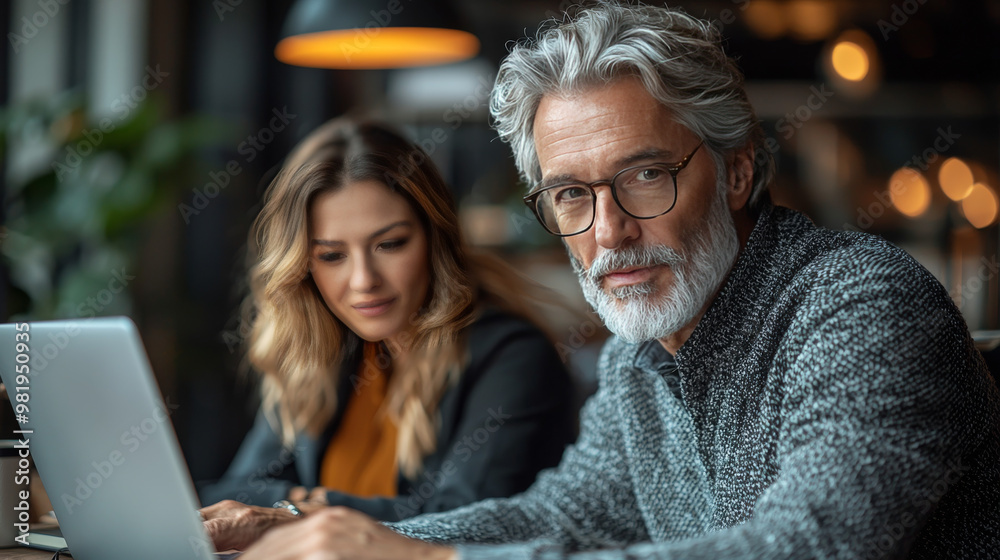 A man with a white beard looks directly at the camera as his female colleague works on a laptop. A man with a white beard looks directly at the camera as his female colleague works on a laptop.
