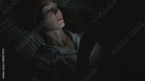 A young woman in bed uses her smartphone to surf the Internet at night. The light from the phone screen