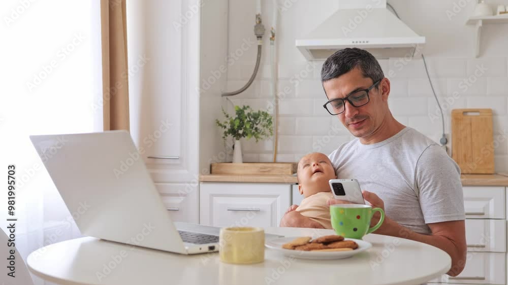 Satisfied cheerful man father talking on smartphone holding baby in kitchen managing business call while working from laptop enjoying time with his child and freelance activity Satisfied cheerful man father talking on smartphone holding baby in kitchen managing business call while working from laptop enjoying time with his child and freelance activity