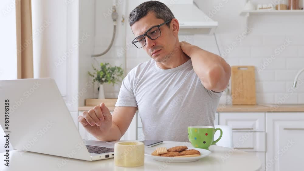 Tired man engaged in digital work and online education at kitchen table feeling tired and massaging neck due to pain after sitting in uncomfortable pose long hours working Tired man engaged in digital work and online education at kitchen table feeling tired and massaging neck due to pain after sitting in uncomfortable pose long hours working