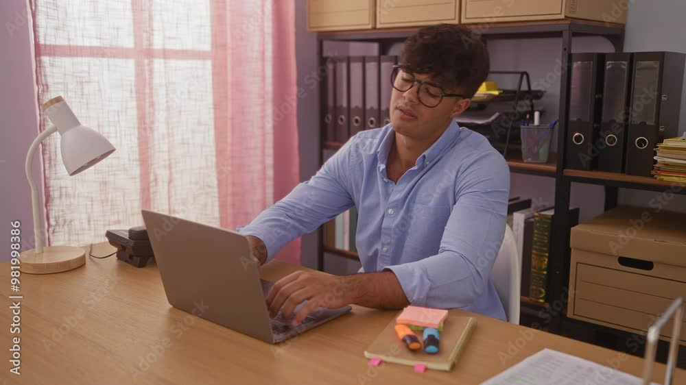 Young hispanic man in an office room closing his laptop and relaxing in a chair after working, captured in a bright, well-organized indoor workplace.