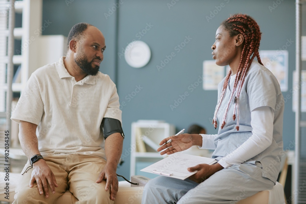 Obraz premium African American male patient sitting on hospital bed engaging with nurse while holding clipboard in medical office setup, displaying focused expressions and gestures