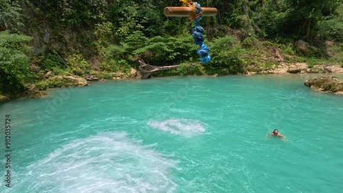 A man swings into bright blue water while canyoneering at Kawasan Falls in Cebu, Philippines. he jumps and swings from a platform and then falls into the water below where a woman is swimming.