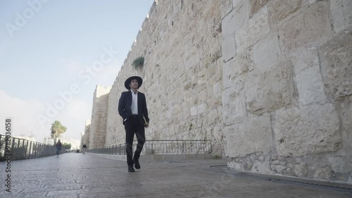 An Orthodox male in traditional clothing walks with purpose along the ancient stone walls of the Old City of David in Jerusalem, reflecting the city's deep history, culture, and spirituality photo