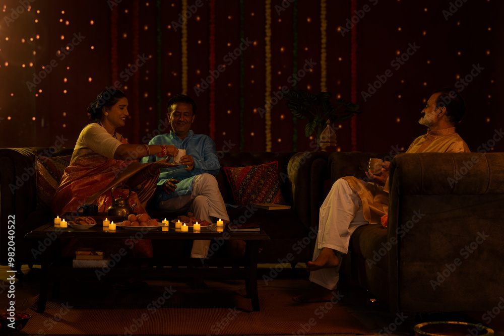 Senior woman offering tea to her husband while sitting together on sofa with their friend at home during Diwali celebration Senior woman offering tea to her husband while sitting together on sofa with their friend at home during Diwali celebration