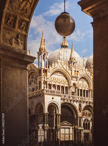 St. Mark’s Basilica in Venice, viewed through a stone arch. The background showcases the building’s rich architecture with its characteristic domes, and a crowd of tourists gathers below on the square photo