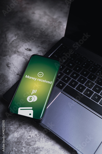Smartphone laying on laptop with money received notification on screen. Selective focus. Concept of financial transactions with mobile devices. Smartphone screen displaying money transfer confirmation