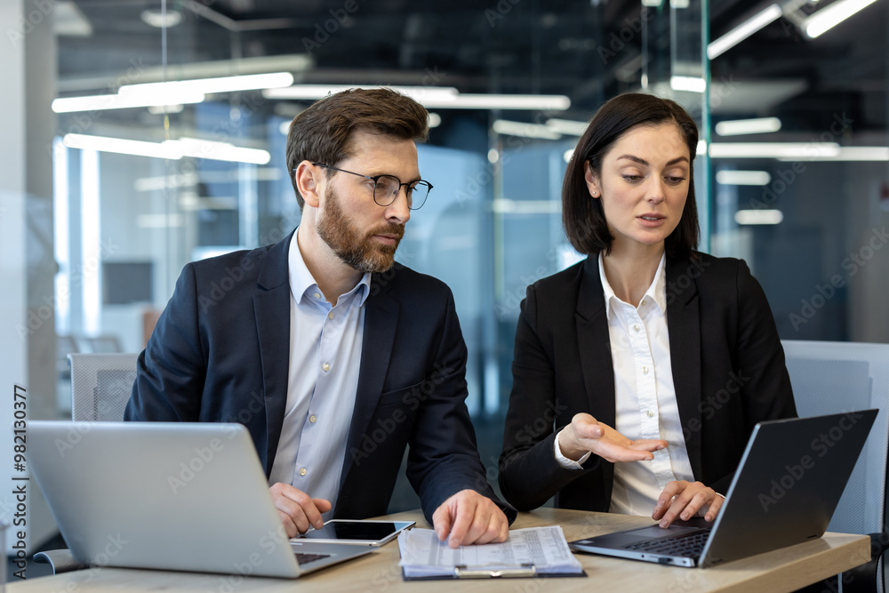 Two colleagues in suits engaged in serious business discussion at office desk. Open laptops and clipboard suggest collaborative work. Focused expressions indicate detailed conversation Two colleagues in suits engaged in serious business discussion at office desk. Open laptops and clipboard suggest collaborative work. Focused expressions indicate detailed conversation