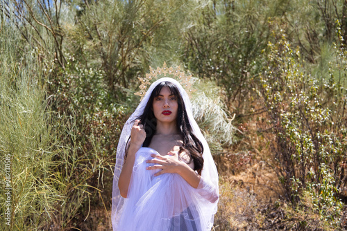 beautiful young brunette latin woman with red lips and dressed with a white veil and a golden crown of virgin mary or goddess. The woman is in a forest with vegetation as if she were an apparition. photo