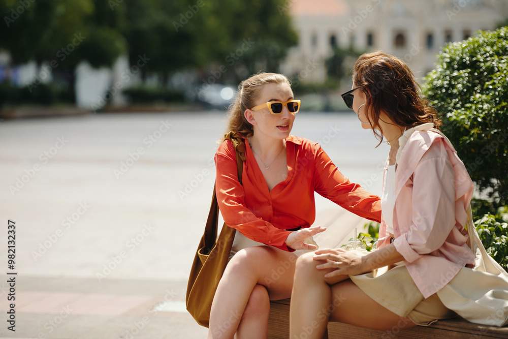 Naklejka premium Two women sitting on park bench and having an animated conversation on sunny day, both wearing casual attire blond-haired woman wearing sunglasses