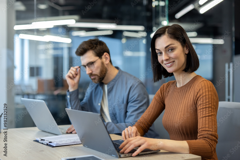 Two business professionals collaborating in a modern office environment. Woman smiling while typing on laptop, man in background working on laptop with thoughtful expression Two business professionals collaborating in a modern office environment. Woman smiling while typing on laptop, man in background working on laptop with thoughtful expression