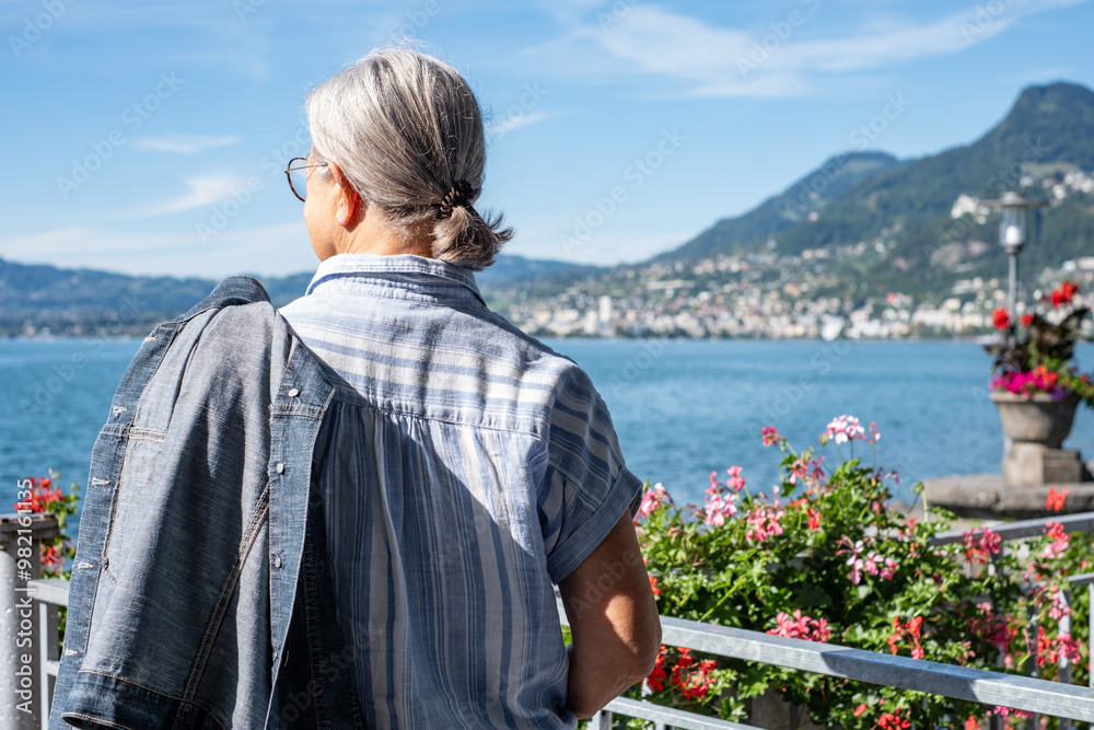 Fototapeta premium Rear view of senior gray haired woman standing along lake admiring the landscape - relaxed elderly grandmother enjoying peaceful moments in vacation, Lake Geneva and mountains in background