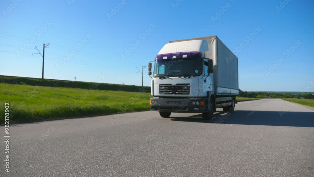 Camera follows to truck with cargo trailer driving on empty highway transporting goods at summer day. White lorry riding through countryside with nature landscape at background. Slow motion Front view
