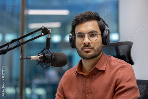 Serious male broadcaster with glasses and headphones sits in studio recording podcast. Professional setting conveys focus and expertise. Capturing essence of modern media and communication.