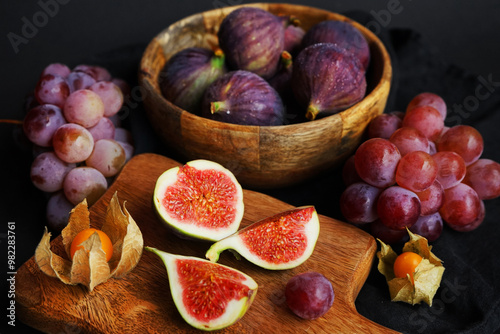 Ripe whole and sliced ​​figs next to pink grapes and Physalis fruits on a wooden board on a dark background