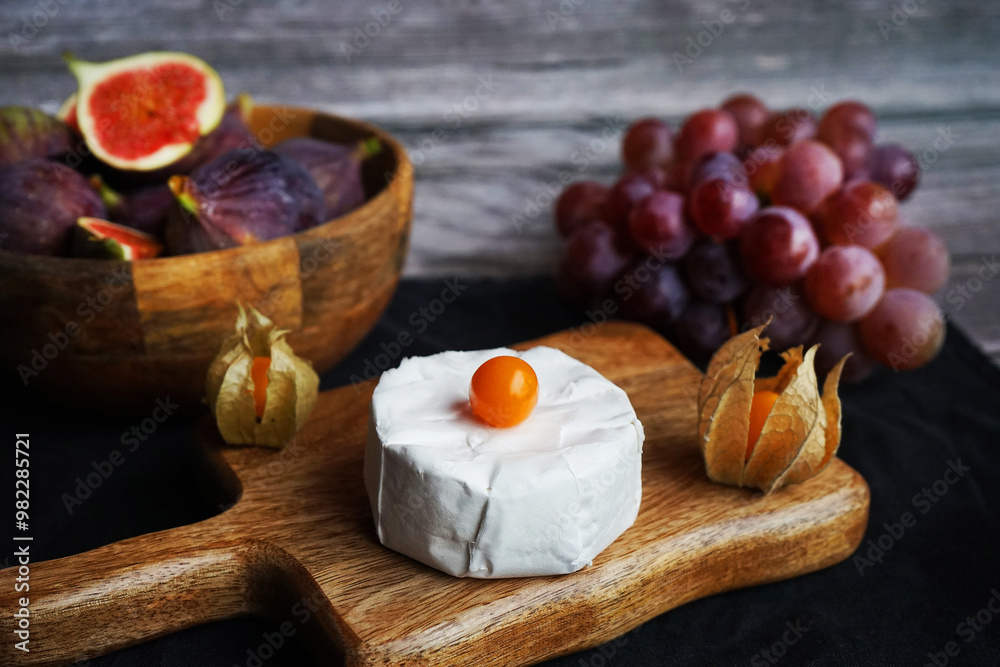 Fototapeta premium Ripe figs next to Brie cheese on a wooden board, pink grapes and Physalis fruits on a dark background
