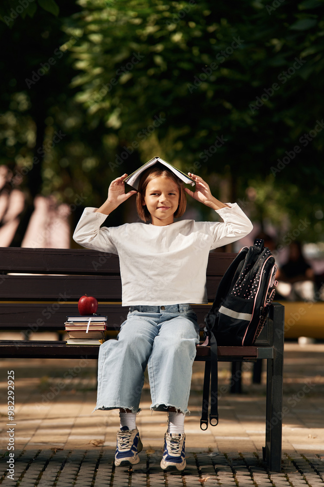 Having fun, book over the head. Schoolgirl is sitting on the bench with a bunch of books outdoors Having fun, book over the head. Schoolgirl is sitting on the bench with a bunch of books outdoors