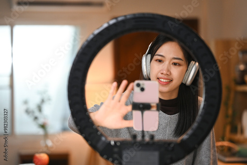 Young woman wearing headphones live streaming or recording content with a smartphone on a tripod stand with a ring light