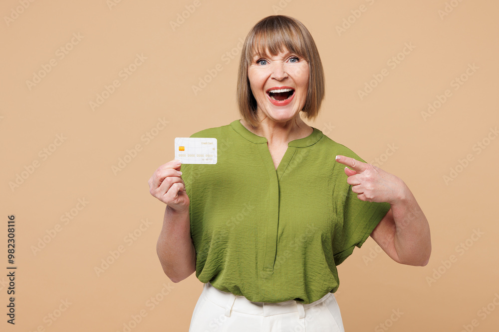 Elderly blonde woman 50s years old wear green shirt casual clothes hold in hand point finger on mock up of credit bank card isolated on plain pastel light beige background studio. Lifestyle concept. Elderly blonde woman 50s years old wear green shirt casual clothes hold in hand point finger on mock up of credit bank card isolated on plain pastel light beige background studio. Lifestyle concept.