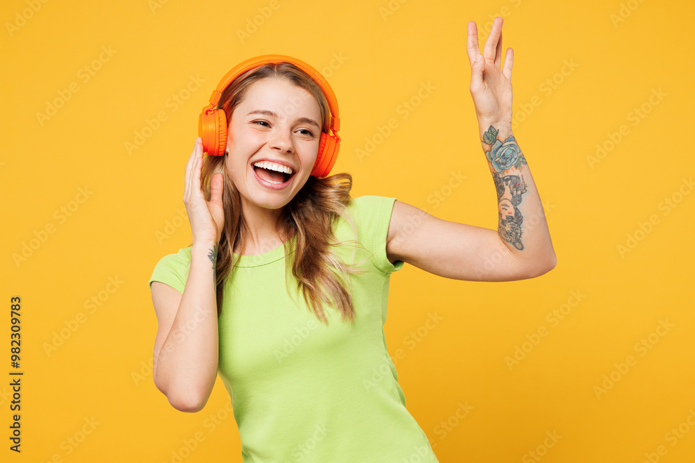 Young happy funny blonde woman she wear green t-shirt casual clothes listen to music in headphones raise up hands dance isolated on plain yellow orange background studio portrait. Lifestyle concept. Young happy funny blonde woman she wear green t-shirt casual clothes listen to music in headphones raise up hands dance isolated on plain yellow orange background studio portrait. Lifestyle concept.