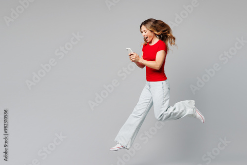 Full body young smiling happy fun woman she wear red t-shirt casual clothes jump high hold in hand use mobile cell phone run fast isolated on plain grey background studio portrait. Lifestyle concept. photo