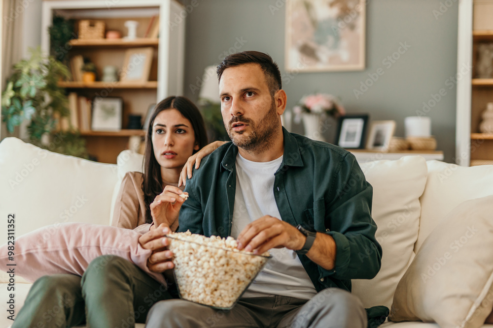 Multiracial husband and wife eating popcorn from a bowl and watching drama movie while chilling on the couch in weekend Multiracial husband and wife eating popcorn from a bowl and watching drama movie while chilling on the couch in weekend