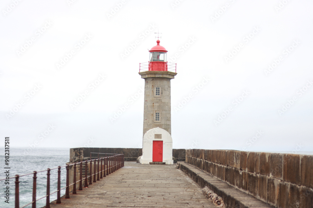 Stone lighthouse with red top and red door on a pier, surrounded by a cloudy sky, located near the coastline on a calm and serene day Stone lighthouse with red top and red door on a pier, surrounded by a cloudy sky, located near the coastline on a calm and serene day