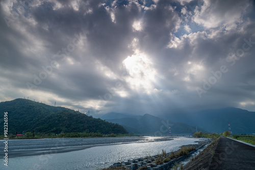 A dramatic interplay of light and shadow unfolds over a river valley. Sunlight intermittently breaks through a canopy of clouds, casting beams of light onto the surrounding mountains. Yilan, Taiwan. photo