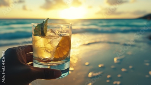 Hand holding a cocktail glass with ice and fruit on the beach at sunset.