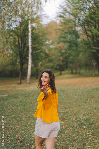 A woman in a bright yellow top walks through a park, smiling happily as she enjoys the beauty of nature and the warmth of the sun in autumn. #982427123