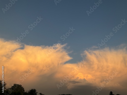 Blue sky with stormy menacing Anvil cloud, Cumulonimbus incus illuminated in orange by sunlight at the evening over dark horizon with trees. Topics: storm alert, meteorology, weather, nature, climate photo