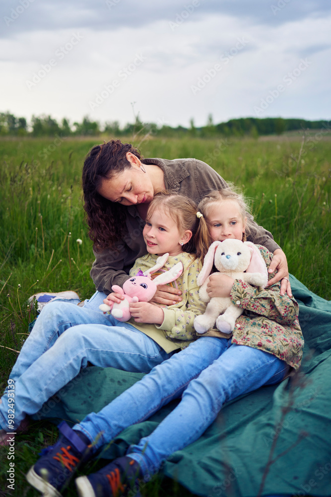 a mother with two children and their toys hug while sitting on a blanket in a field a mother with two children and their toys hug while sitting on a blanket in a field