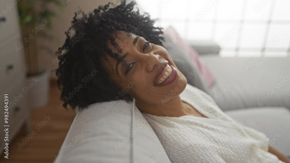 A beautiful young african american woman with curly hair smiles warmly while relaxing on a sofa in a cozy, well-lit living room. A beautiful young african american woman with curly hair smiles warmly while relaxing on a sofa in a cozy, well-lit living room.