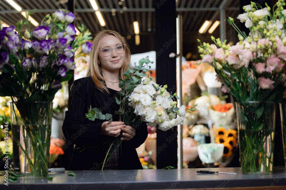 Fototapeta premium Happy young woman florist in suit collecting bouquet at workplace in flower shop