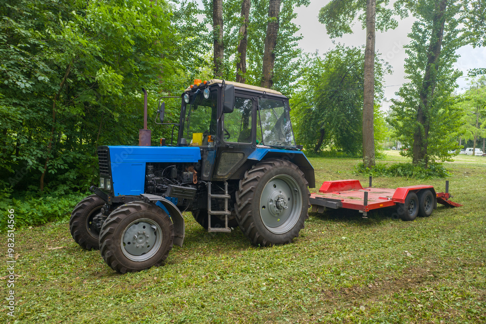 A blue tractor is parked in a field next to a red trailer. The tractor is surrounded by trees and grass A blue tractor is parked in a field next to a red trailer. The tractor is surrounded by trees and grass