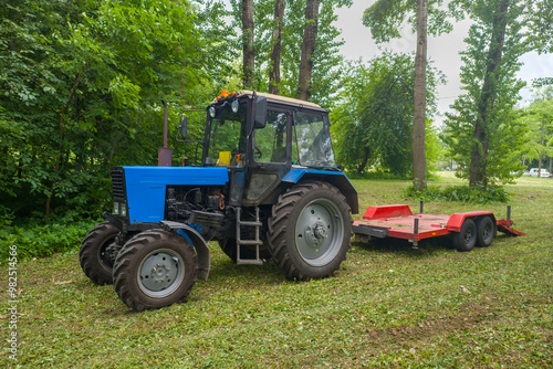 A blue tractor is parked in a field next to a red trailer. The tractor is surrounded by trees and grass