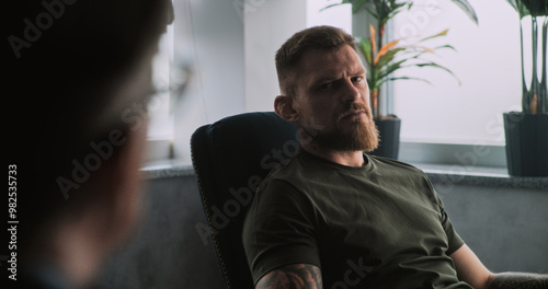 Depressed military man in camouflage with PTSD sits in chair, shares feelings with psychotherapist or psychologist on rehabilitation therapy session. Post traumatic stress disorder. Over the shoulder.