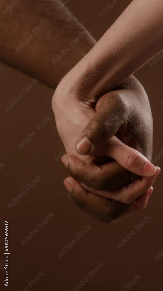 Vertical detail shot of diverse romantic couple slowly interlocking fingers against brown background Vertical detail shot of diverse romantic couple slowly interlocking fingers against brown background