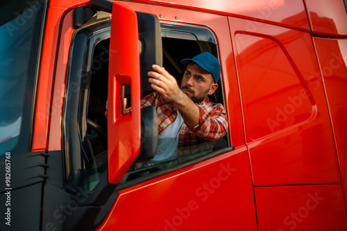 Caucasian truck driver adjusting the side mirror of a bright red truck.