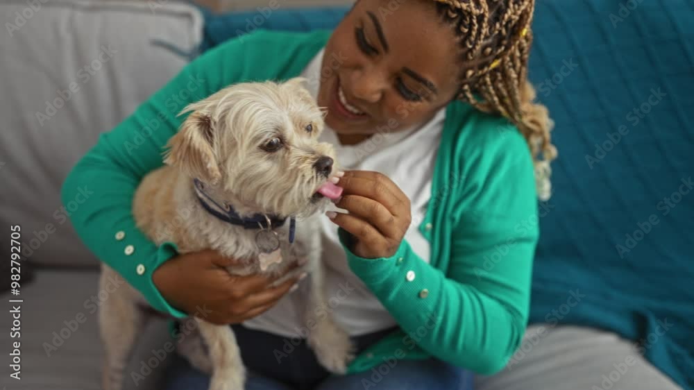 A young african american woman with braids, wearing a green cardigan, plays with her dog on the couch in a cozy living room.