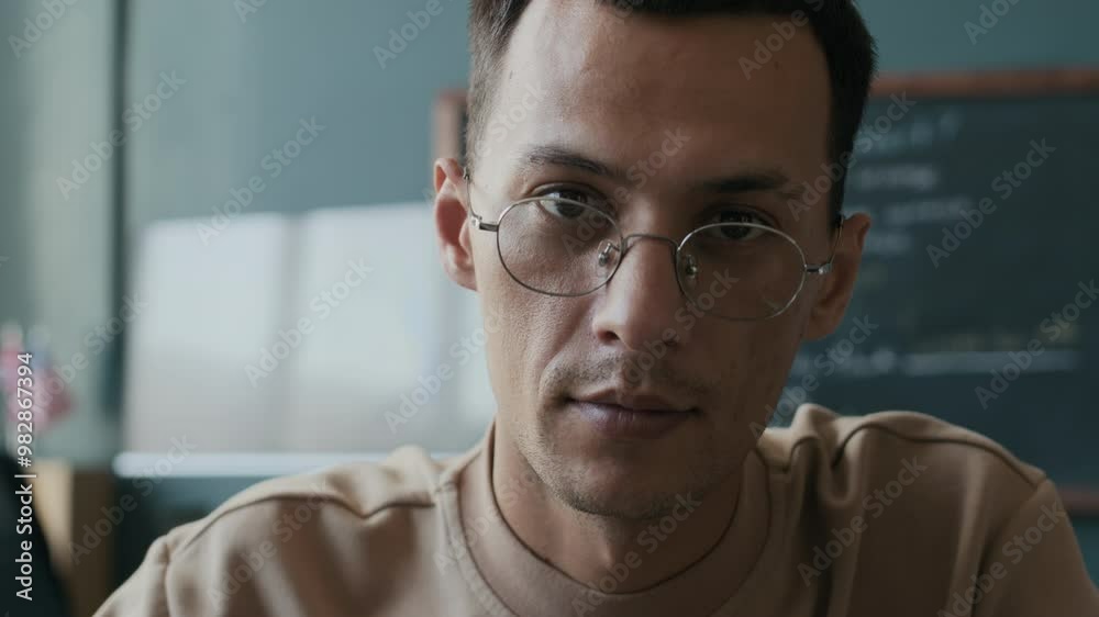 Zoom-in closeup portrait of young adult male university student in glasses sitting at desk in classroom and completing worksheet Zoom-in closeup portrait of young adult male university student in glasses sitting at desk in classroom and completing worksheet