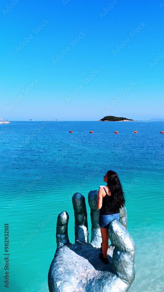 Long-haired brunette standing at the sculpture of hand on the sandy beach. Drone footage above the blue waterscape approaching red buoys. Vertical video. Long-haired brunette standing at the sculpture of hand on the sandy beach. Drone footage above the blue waterscape approaching red buoys. Vertical video.