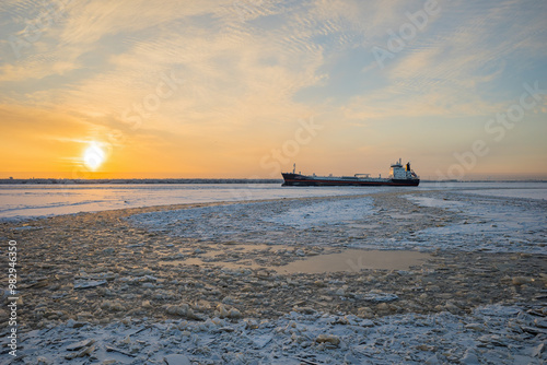 in winter, in severe frost, a cargo ship passes along the breaks made in the frozen Severnaya Dvina River in the city of Arkhangelsk, against the background of the setting sun