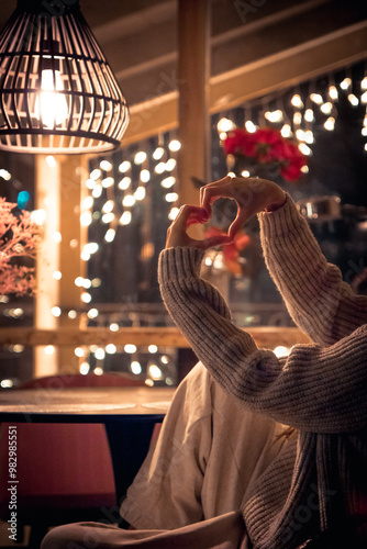 Hands heart and love symbol made by woman sitting in warm house in cold winter night with fairy lights on background