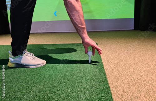 A golfer positions a golf ball on the artificial turf in an indoor facility, focusing on his grip