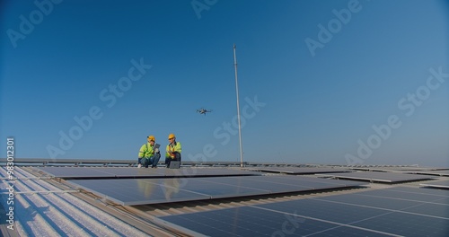 Two engineers with safety vests and hard hats are inspecting solar panels with a drone flying overhead against a clear blue sky