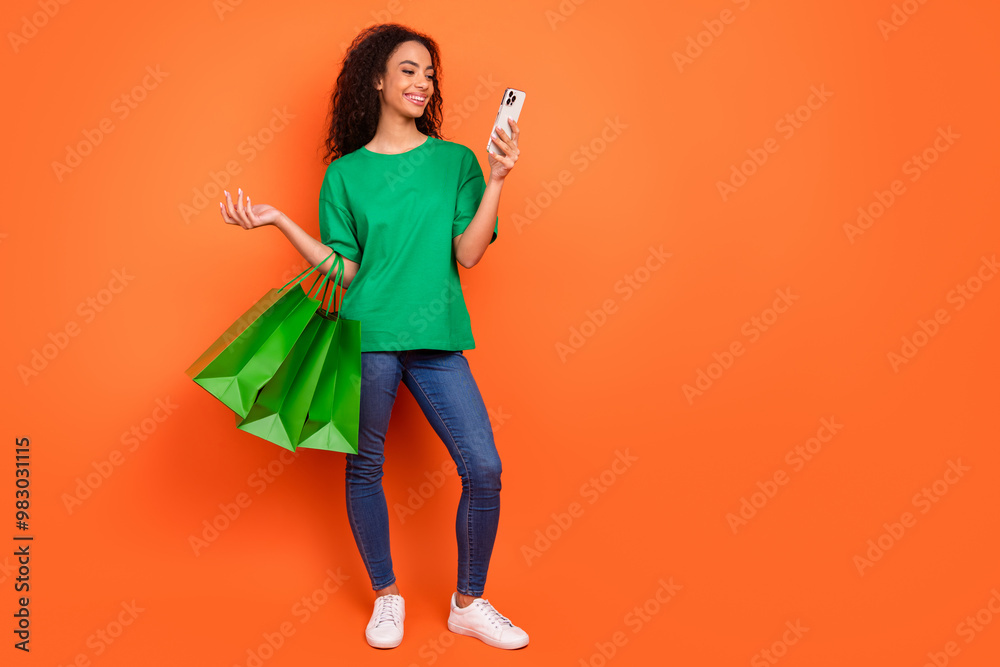 Photo of young attractive girl wear green casual t shirt and jeans holding shopping bags surfing wish list isolated on orange color background Photo of young attractive girl wear green casual t shirt and jeans holding shopping bags surfing wish list isolated on orange color background