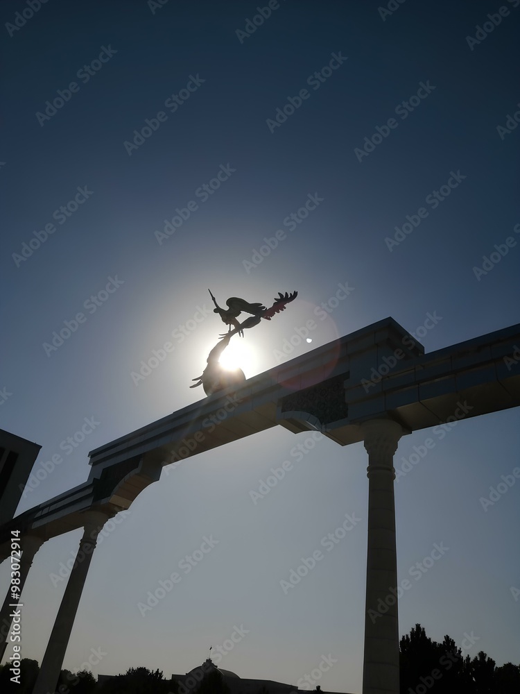 The sun is setting in the background, with a bright sun flare, and a silhouette of a sculpture of two birds on a building's archway. The sun is setting in the background, with a bright sun flare, and a silhouette of a sculpture of two birds on a building's archway.
