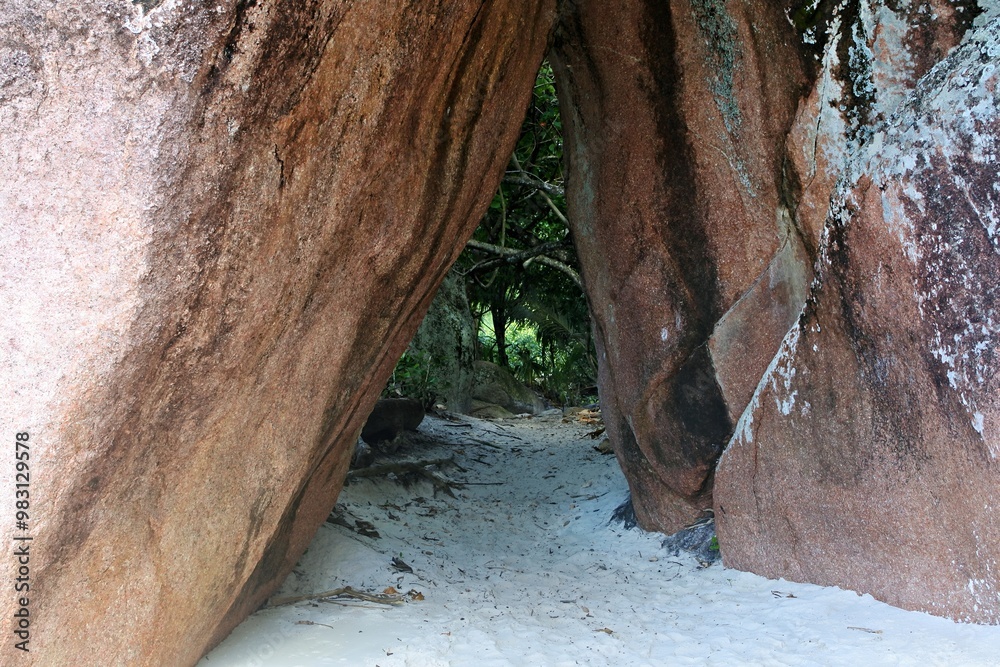 Obraz premium A narrow passageway between large rocks leading to a sandy area surrounded by greenery.